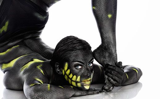 Eine Frau liegt mit der Brust auf dem Bauch, ihr Körper ist mit einem Bodypainting in dunklen Farben und Neon-Gelb bedeckt.