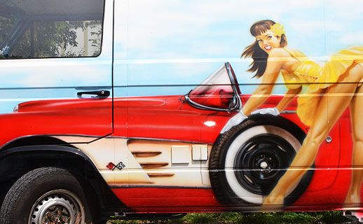 Ein Kleintransporter mit neuer Lackierung, auf der eine Frau neben einem Oldtimer steht.