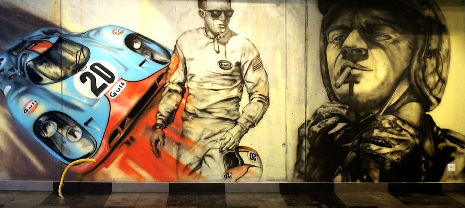 Wandbemalung mit einem blau-roten Rennfahrzeug, einem großen Rennfahrer mit Zigarette und einem weiteren Rennfahrer, der seinen Helm befestigt.